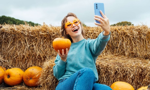Smiling woman in pumpkin-shaped glasses takes a selfie with a pumpkin in front of hay bales.