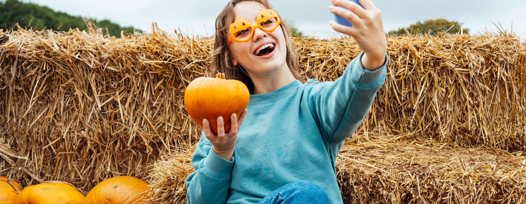 Smiling woman in pumpkin-shaped glasses takes a selfie with a pumpkin in front of hay bales.
